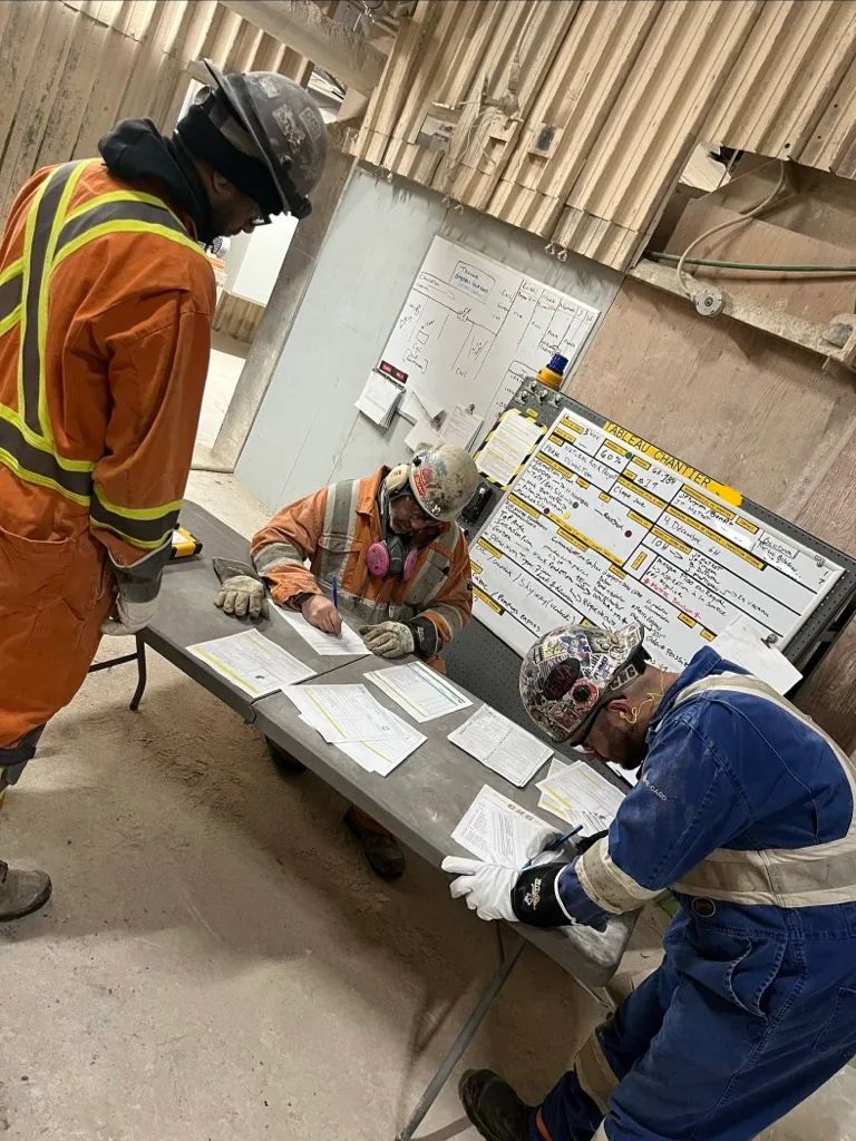 Trois travailleurs en EPI remplissant des formulaires à une table près d’un tableau blanc ou de chantier.