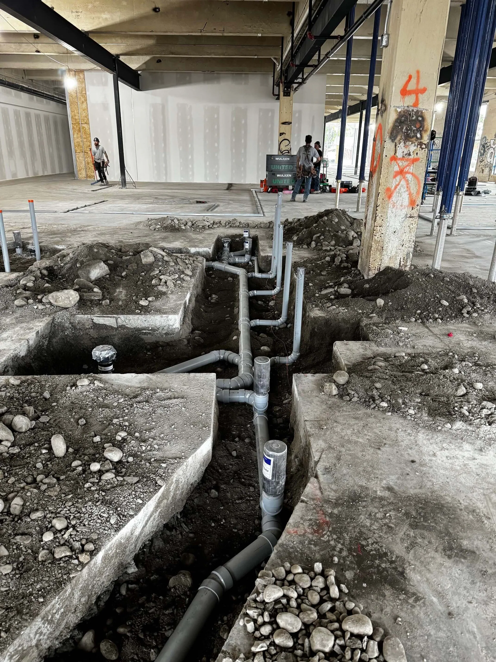 Tuyauterie grise de drainage sous dalle exposée dans des tranchées pendant une rénovation commerciale, avec des travailleurs à l’arrière-plan.