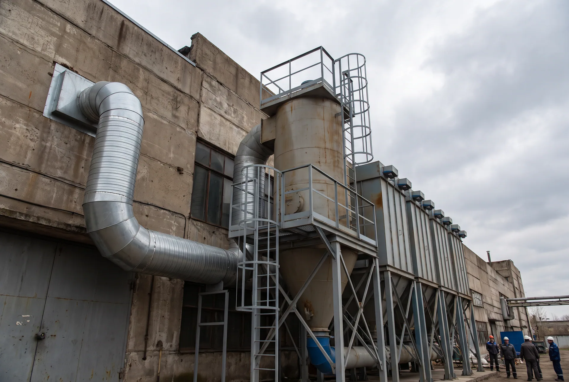 Système de dépoussiérage industriel générique : collecteur à manches, trémie et conduits spiralés le long d’un bâtiment, sans client ni installation identifiable.