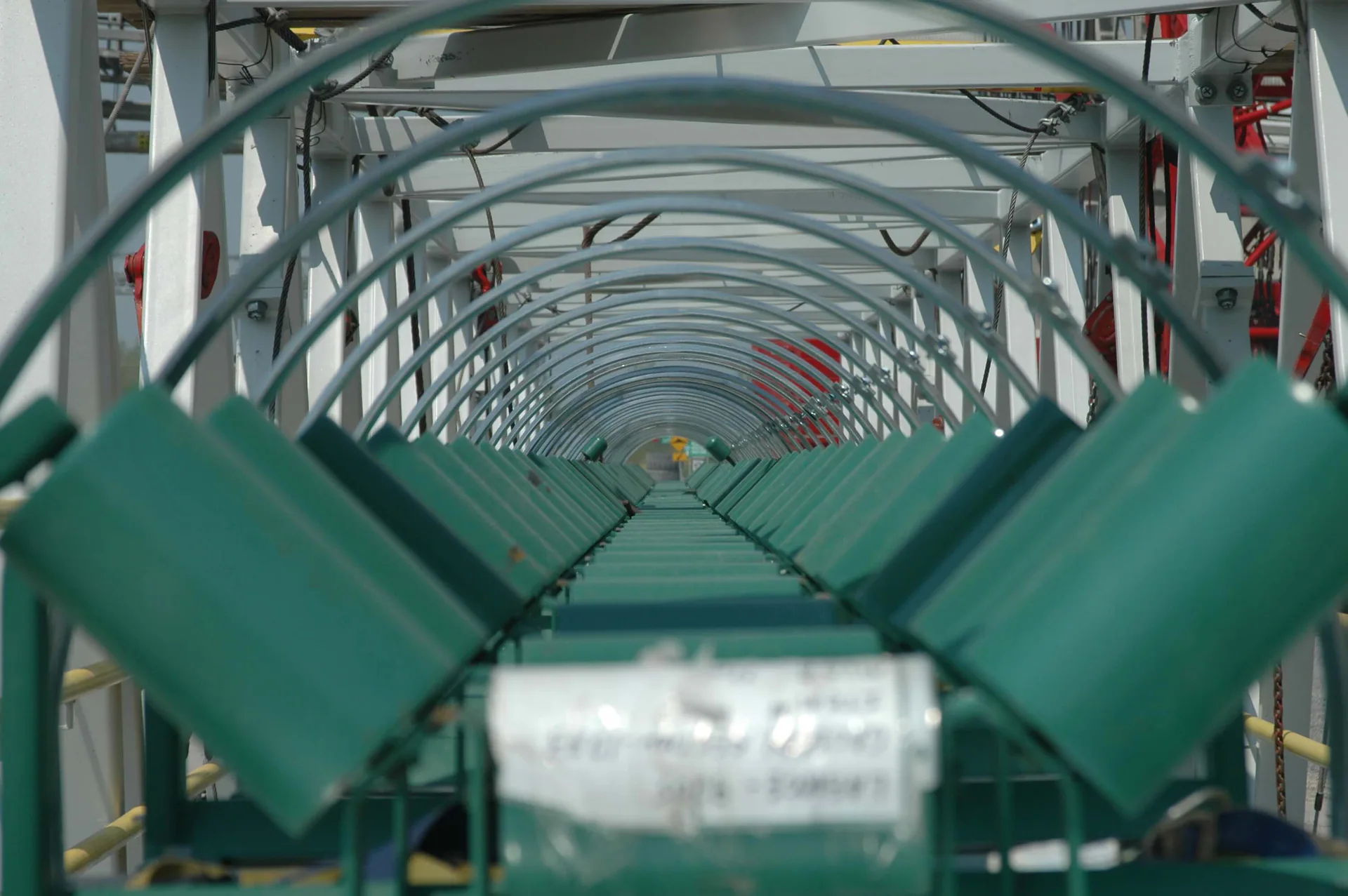 Vue rapprochée le long de rouleaux porteurs verts de convoyeur sous une série de protecteurs arqués.