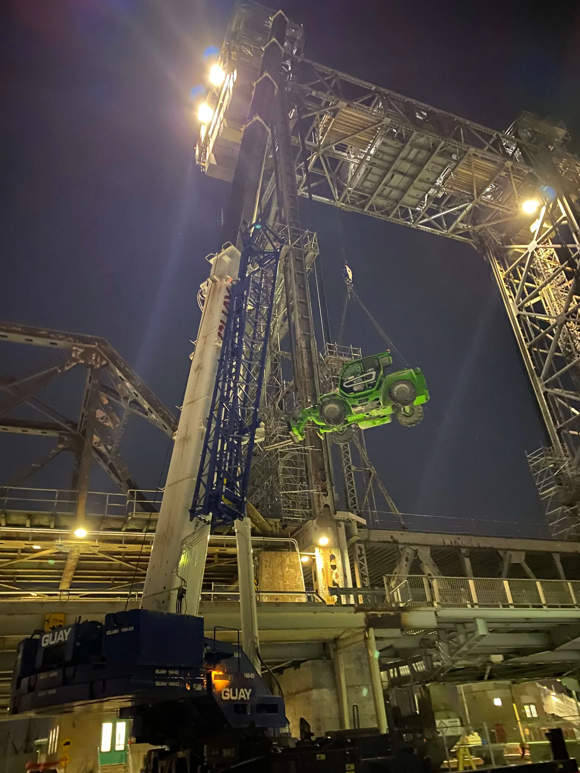 Levage nocturne spectaculaire d’une grande machine mobile verte suspendue dans une haute structure d’acier industrielle, avec grues et projecteurs.