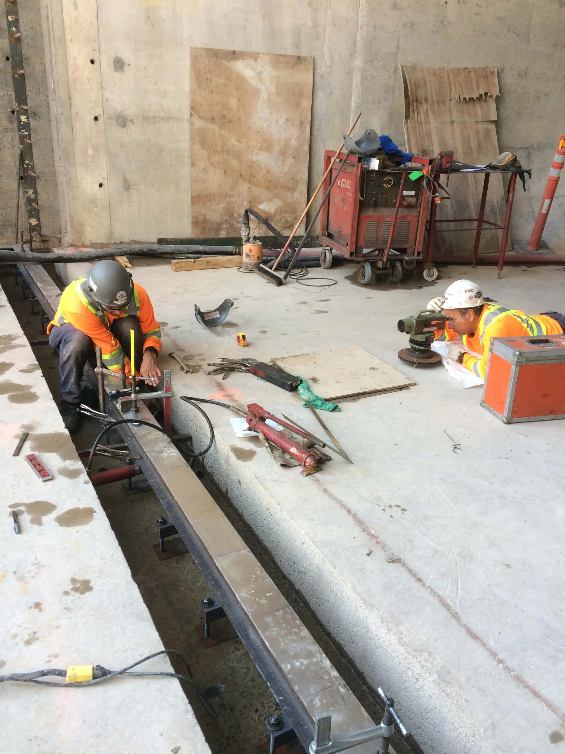 Deux travailleurs en EPI portant la marque CNC mesurant et positionnant un long rail ou profilé d’acier près d’une ouverture de plancher à l’aide d’un instrument d’arpentage.
