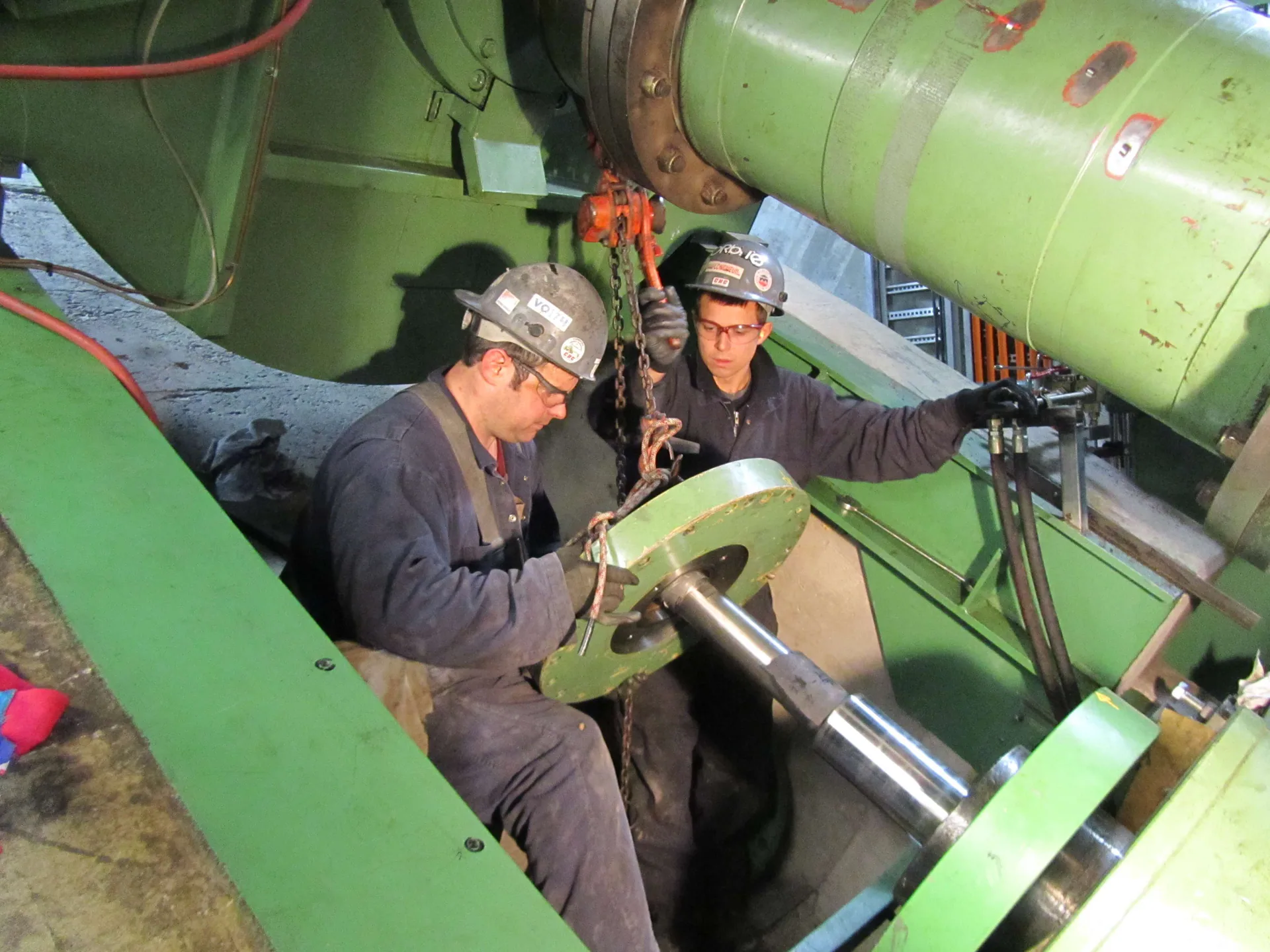 Deux travailleurs casqués manutentionnant un composant d’arbre ou de palier avec un palan à chaîne à l’intérieur d’une grande machine rotative verte.