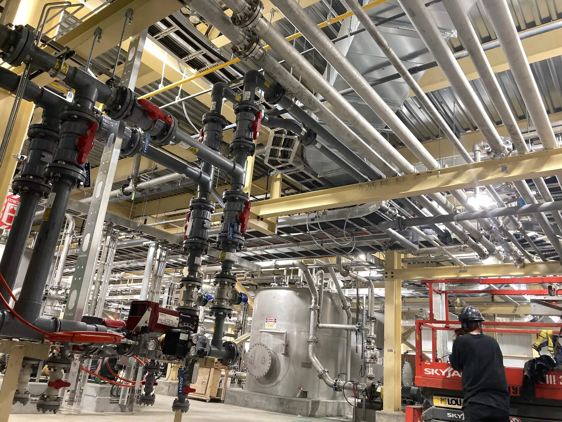 Réseau intérieur dense de tuyauterie de procédé comprenant conduites en acier inoxydable, vannes, réservoirs, supports et un travailleur ou une plateforme élévatrice en bas à droite.