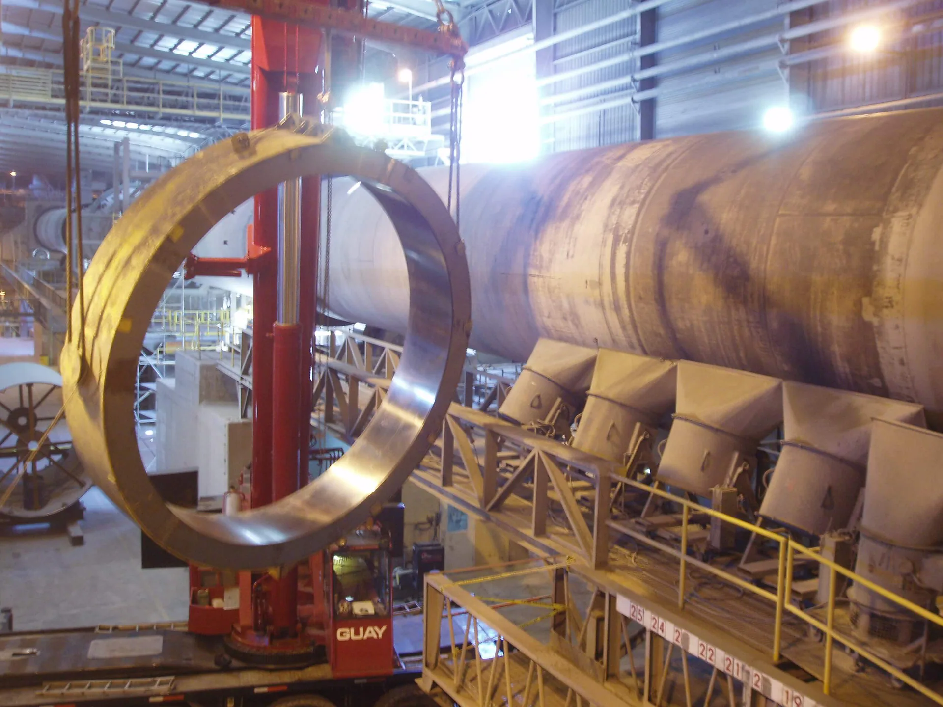 Immense anneau d’acier suspendu à une grue mobile près d’une longue enveloppe cylindrique industrielle.