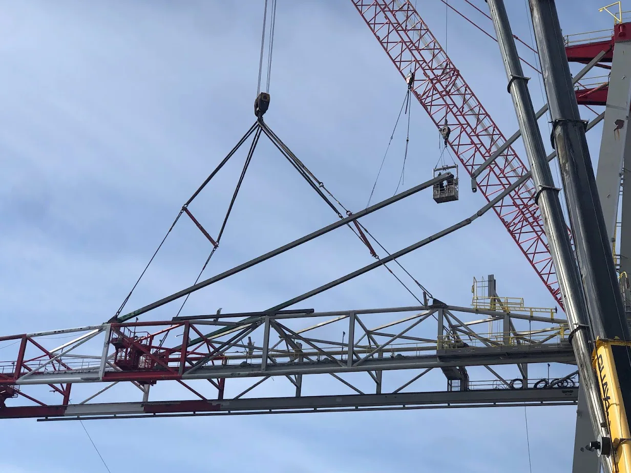 Longue section de treillis ou de flèche industrielle suspendue au gréage d’une grue près d’une machine de type empileuse-récupératrice.