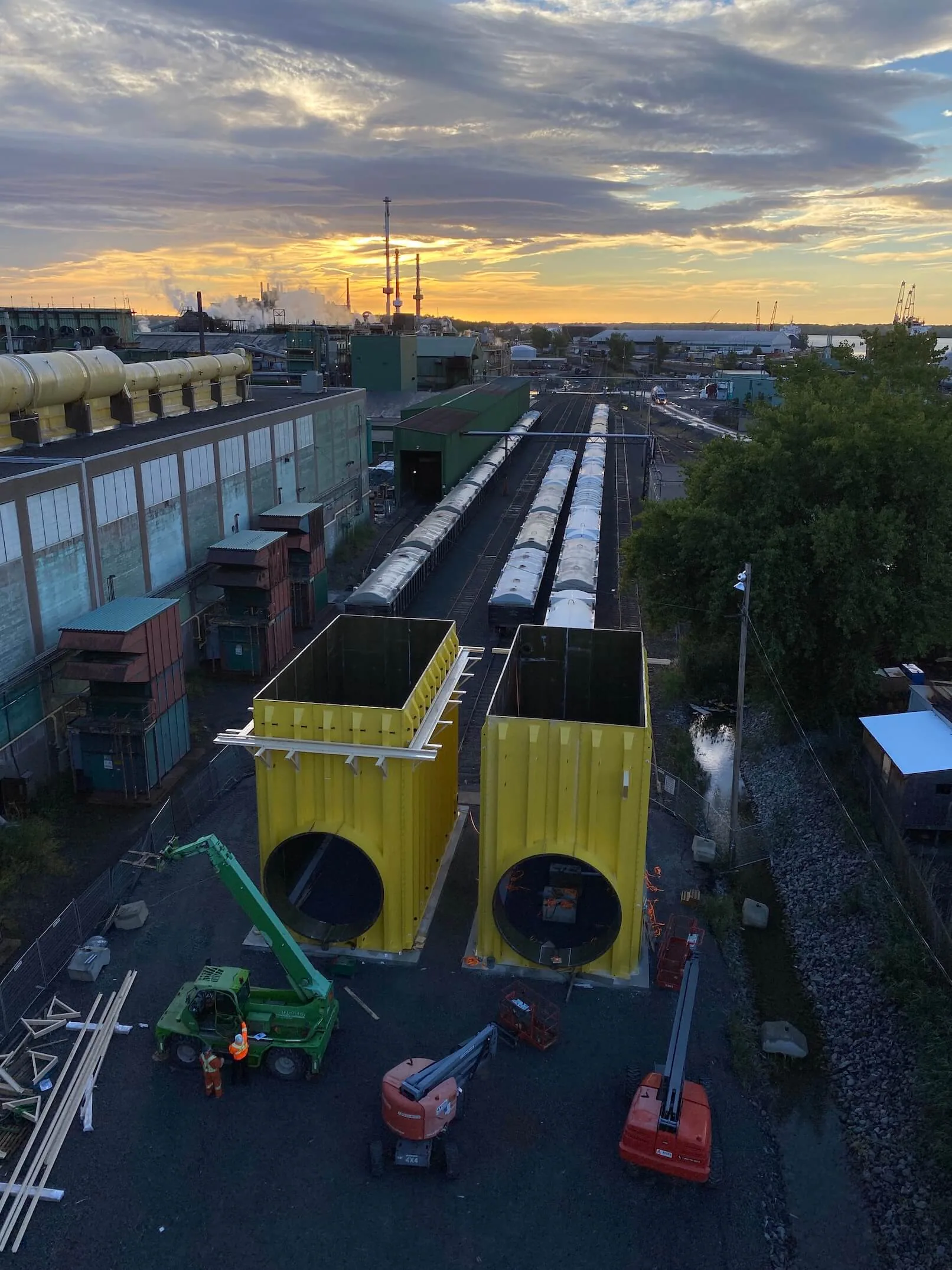 Vue en plongée au coucher du soleil de deux grands modules d’acier jaunes ouverts sur le dessus avec ouvertures circulaires, équipement mobile, wagons et vaste installation industrielle.