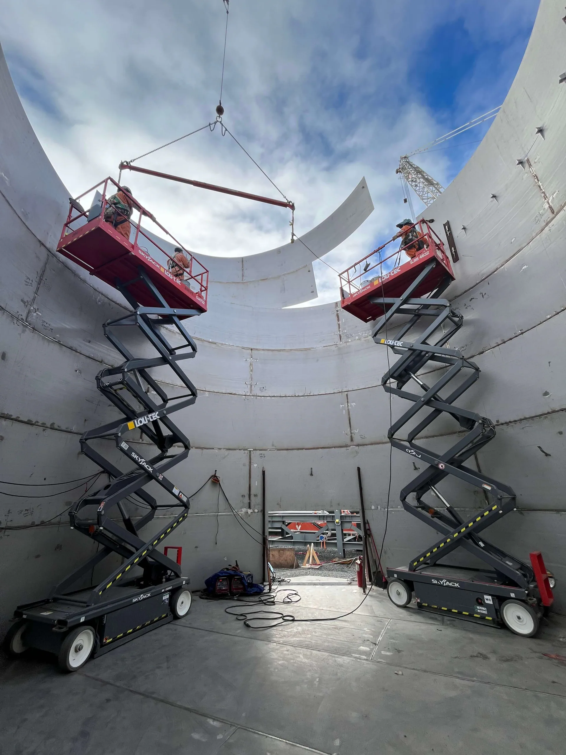 À l’intérieur d’un très grand réservoir circulaire ou d’une enveloppe métallique, deux travailleurs sur plateformes élévatrices guident une plaque d’acier courbée suspendue à une grue ou à un palonnier.