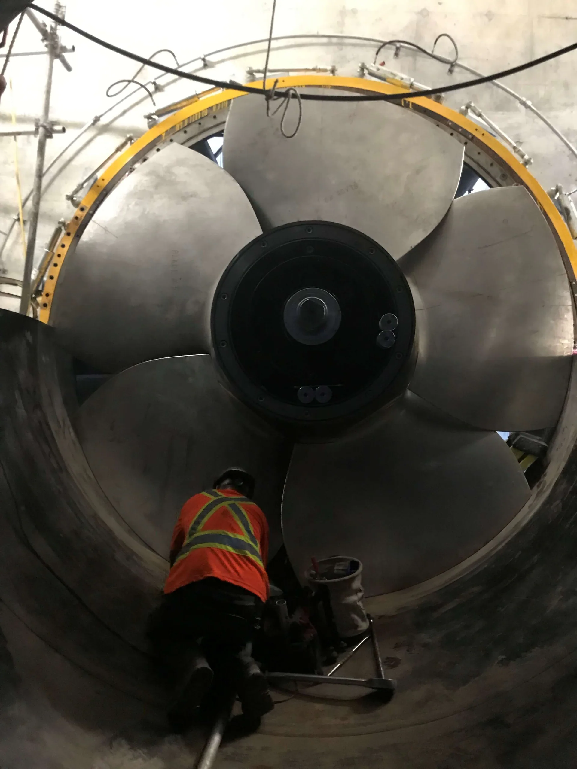 Travailleur à l’intérieur d’un immense carter circulaire sous un énorme assemblage à plusieurs pales de type ventilateur axial, hélice ou roue de turbine.