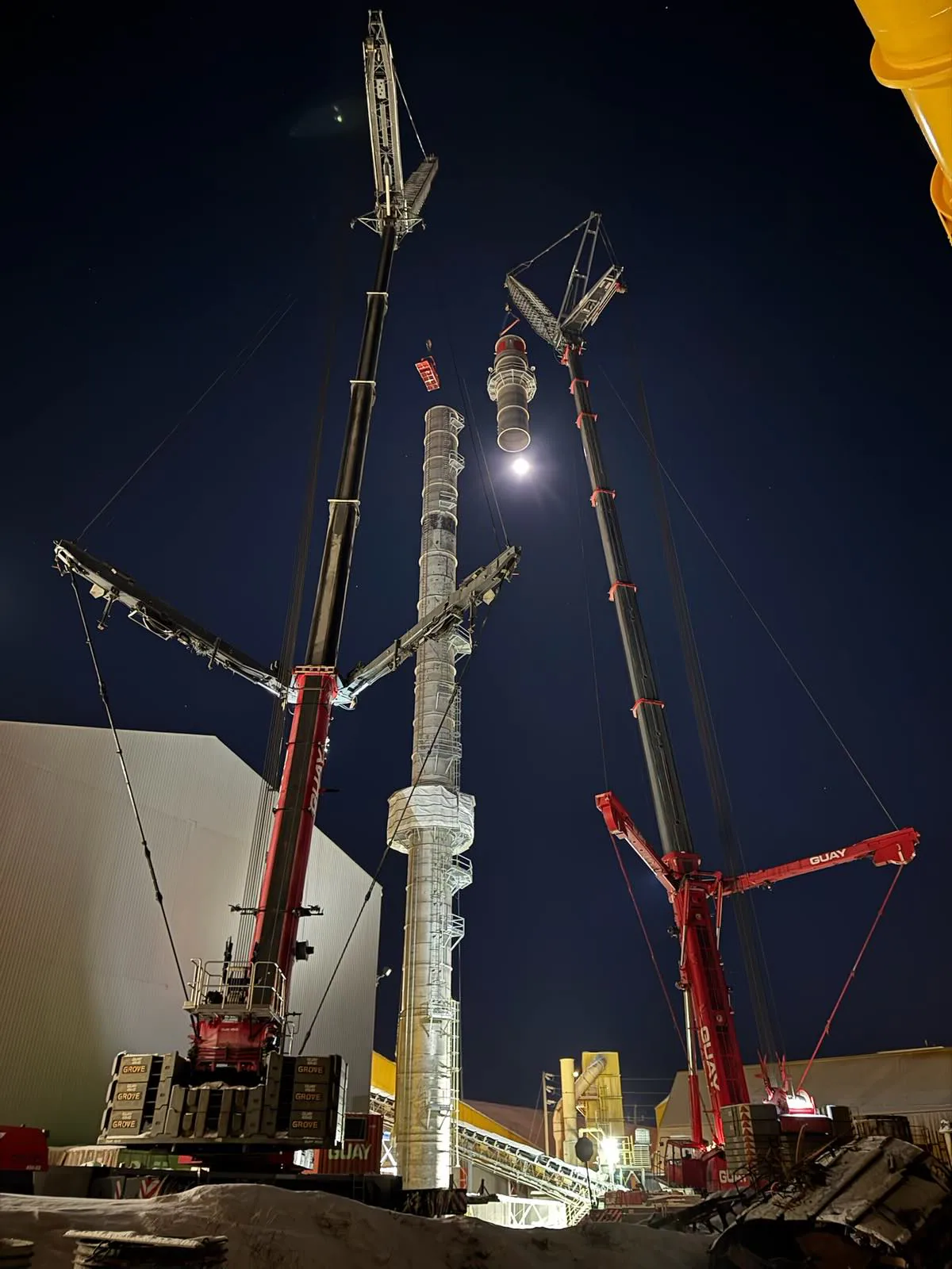 Opération nocturne spectaculaire avec plusieurs grues autour d’une haute section de cheminée ou de colonne industrielle.