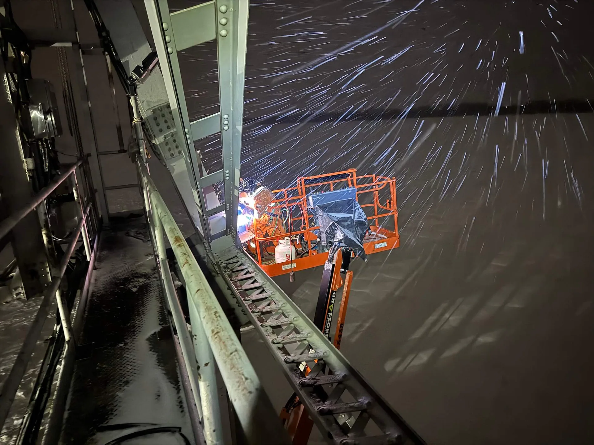 Travailleur dans une nacelle soudant ou découpant un grand élément d’acier de pont la nuit en hiver, avec étincelles visibles.